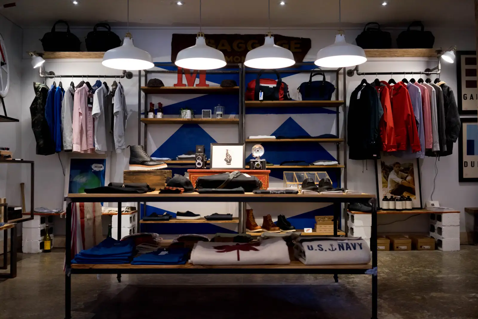 A shop with shelves of folded clothes, bags and shoes under four white pendant lamps
